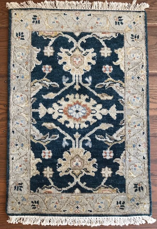 A decorative area rug with a navy blue central pattern featuring floral and geometric motifs, surrounded by an ornate beige border with small intricate designs and a fringed edge.