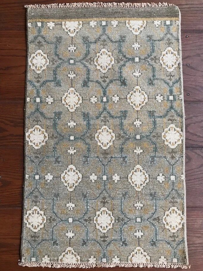 A rectangular area rug with a geometric pattern in shades of beige, gray, and white on a wooden floor.