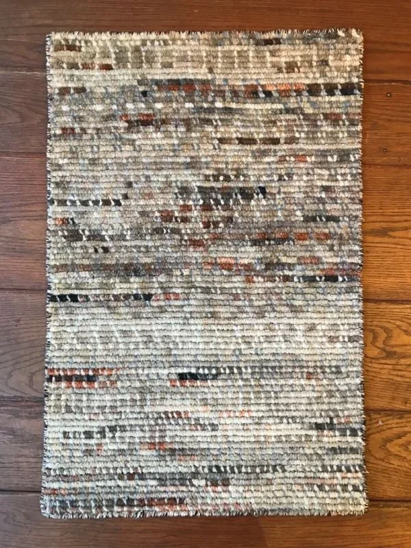 A rectangular area rug with a multicolored, textured pattern in shades of beige, brown, black, and rust on a wooden floor.