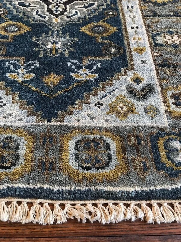 Close-up of a patterned area rug with a floral and geometric design in shades of blue, gold, white, and beige, with fringed edges.