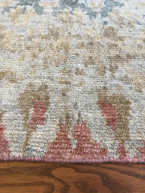 Close-up of a textured area rug with beige, pink, gray, and light blue colors, placed on a wooden floor.
