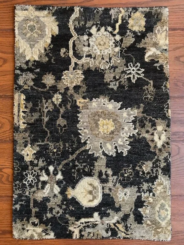 A small, rectangular, patterned area rug with floral and vine designs in black, beige, and cream colors on a wooden floor.