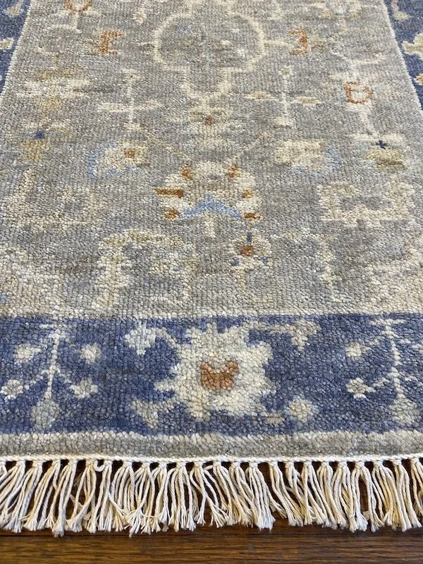 Close-up of a woven rug with a floral and geometric pattern in blue, beige, and brown tones, featuring fringes at the bottom edge.