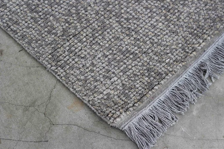 Close-up of a textured gray and beige area rug with fringes, placed on a cracked concrete floor.