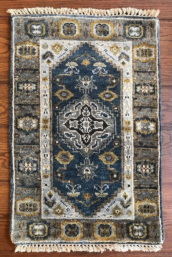 Small decorative rug with a floral and geometric pattern in blue, beige, black, and gold tones on a wooden floor.