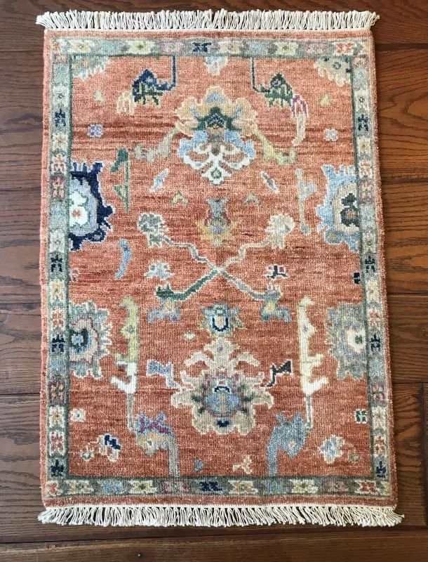 A small, rectangular area rug with a peach background and floral and geometric patterns in shades of blue, green, cream, and black, featuring fringed edges on a wooden floor.