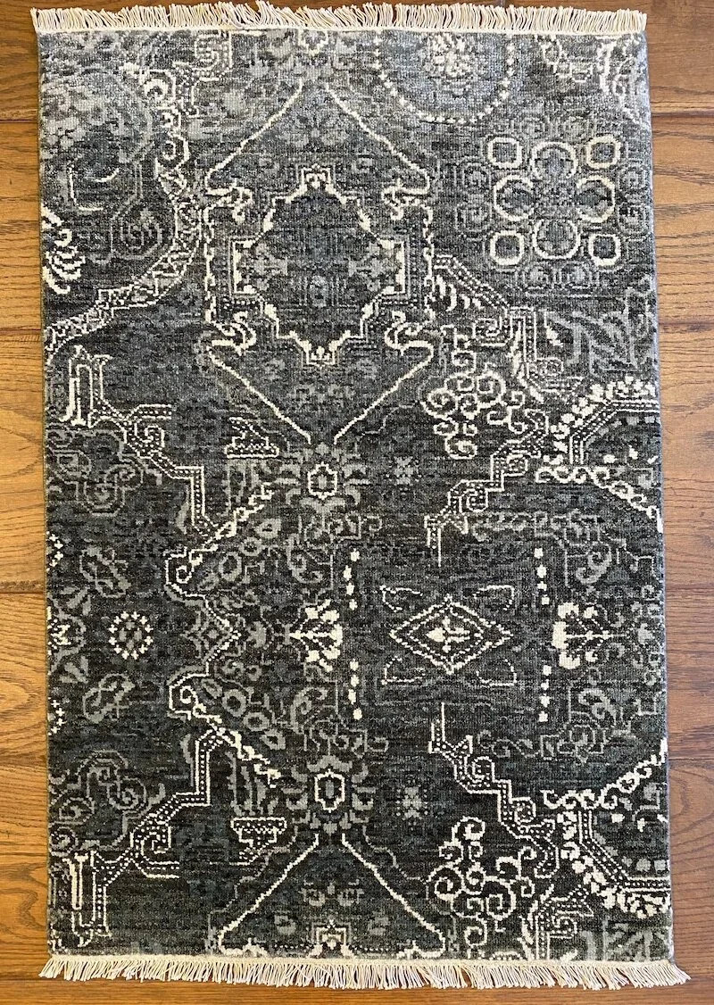 A rectangular area rug with a dark background and intricate light-colored geometric and floral patterns, fringed on the shorter sides, placed on a wooden floor.