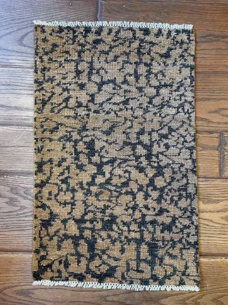Small rectangular patterned rug on hardwood floor, beige and black abstract design with fringed edges.