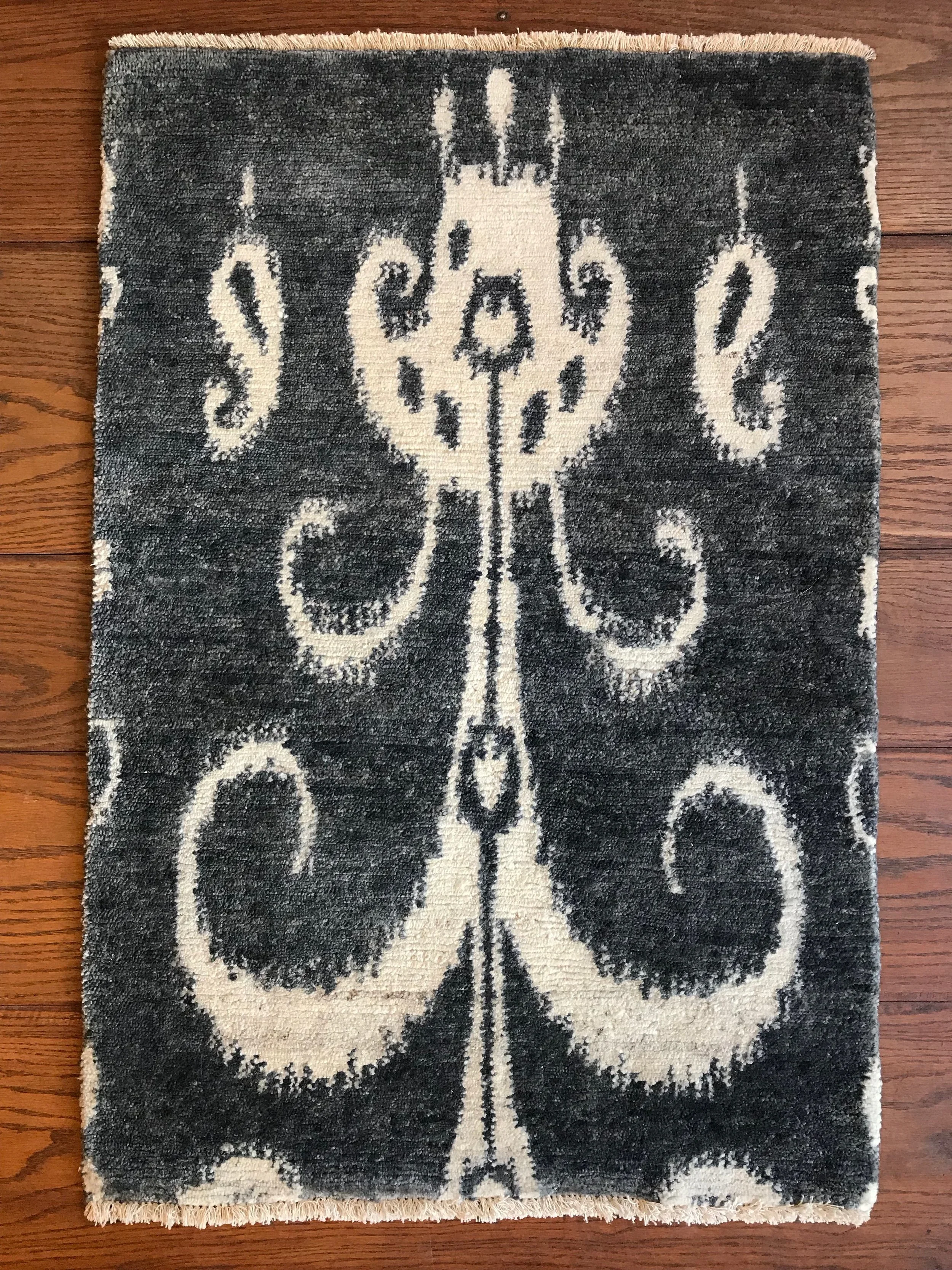 A decorative rug with a black background and a white abstract design resembling a symmetrical pattern with swirling and organic shapes, placed on a wooden floor.