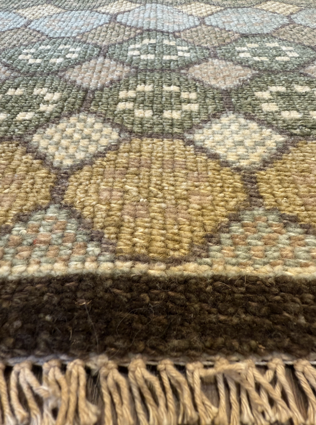 Close-up view of a woven rug with geometric and floral patterns in earthy tones, including green, beige, tan, and brown, and fringes at the edge.