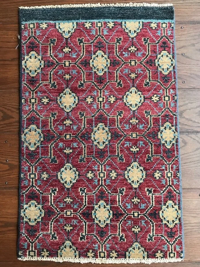 A traditional patterned area rug with a predominantly red background, featuring beige, blue, and black geometric floral motifs and a dark green border along the top edge, placed on a wooden floor.