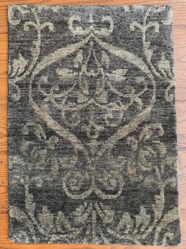 A rectangular rug with a traditional ornate pattern in shades of gray and beige on a wooden floor.