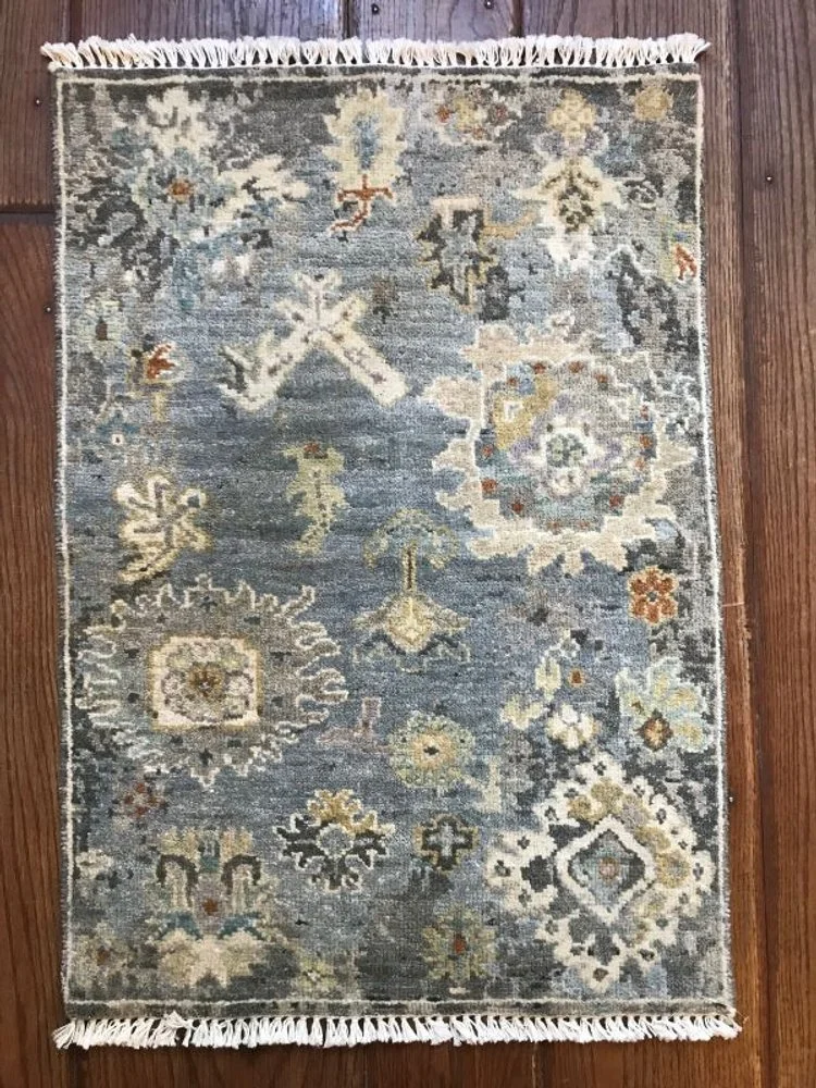 Small decorative rug with a traditional pattern in shades of blue, beige, white, and hints of orange, featuring floral and geometric motifs, fringed edges, placed on a wooden floor.