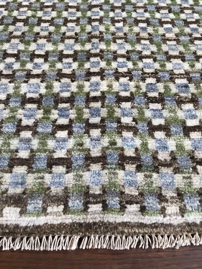 Close-up of a patterned woven rug with a checkered design in blue, white, black, and green colors, hanging with fringe at the bottom.