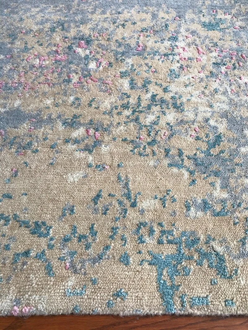 Close-up of a patterned area rug with beige, blue, white, and pink accents on a hardwood floor.