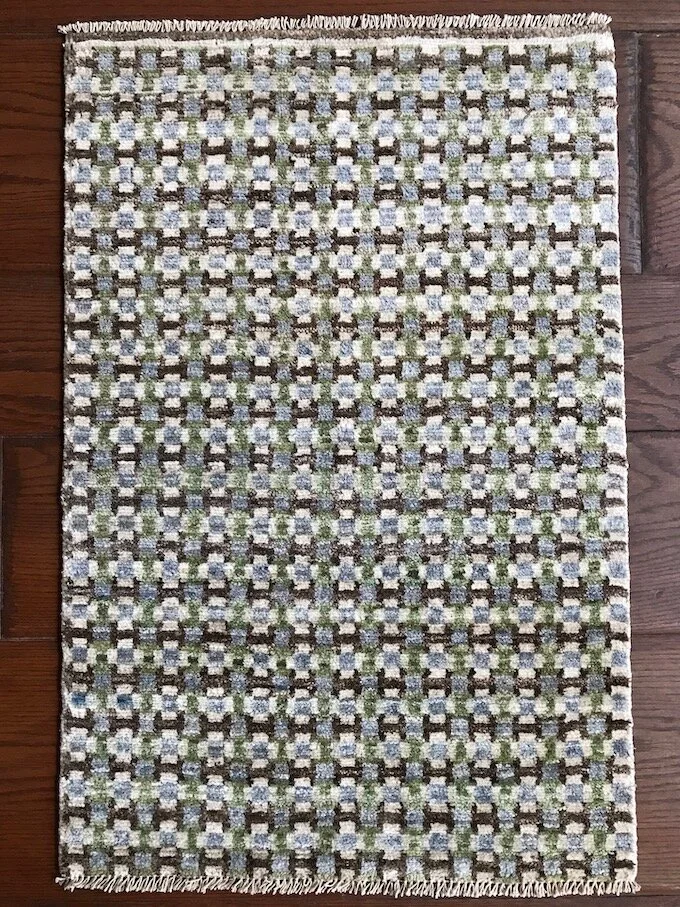 A small rectangular patterned rug with fringe on the top and bottom edges, laid on a wooden floor. The pattern consists of geometric shapes in white, blue, green, and dark brown.