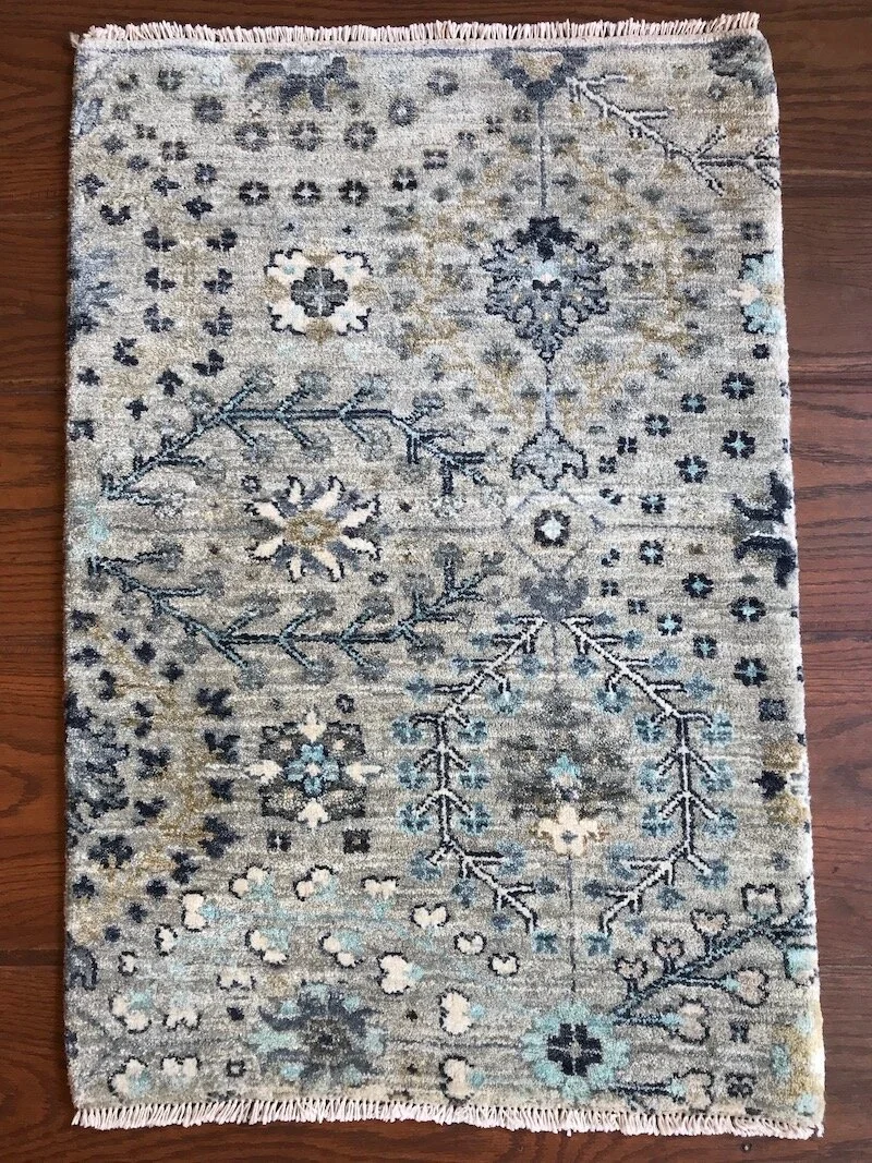 A rectangular decorative area rug with a floral and geometric pattern in shades of beige, gray, blue, and black, placed on a wooden floor.