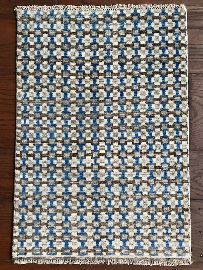 A rectangular woven rug with a geometric pattern in beige, white, blue, and gray colors on a wooden floor.