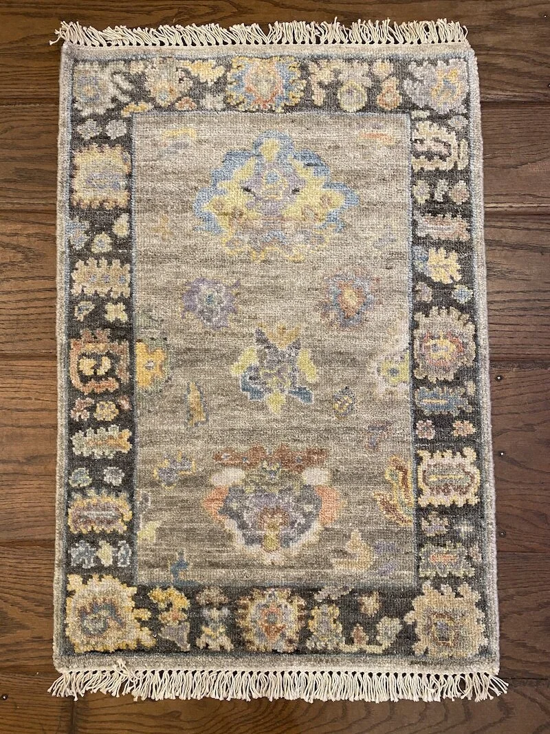 A vintage rectangular rug with a central ornate pattern in pastel colors such as blue, yellow, purple, and orange, bordered by a dark edge with floral and geometric motifs, on a wooden floor.