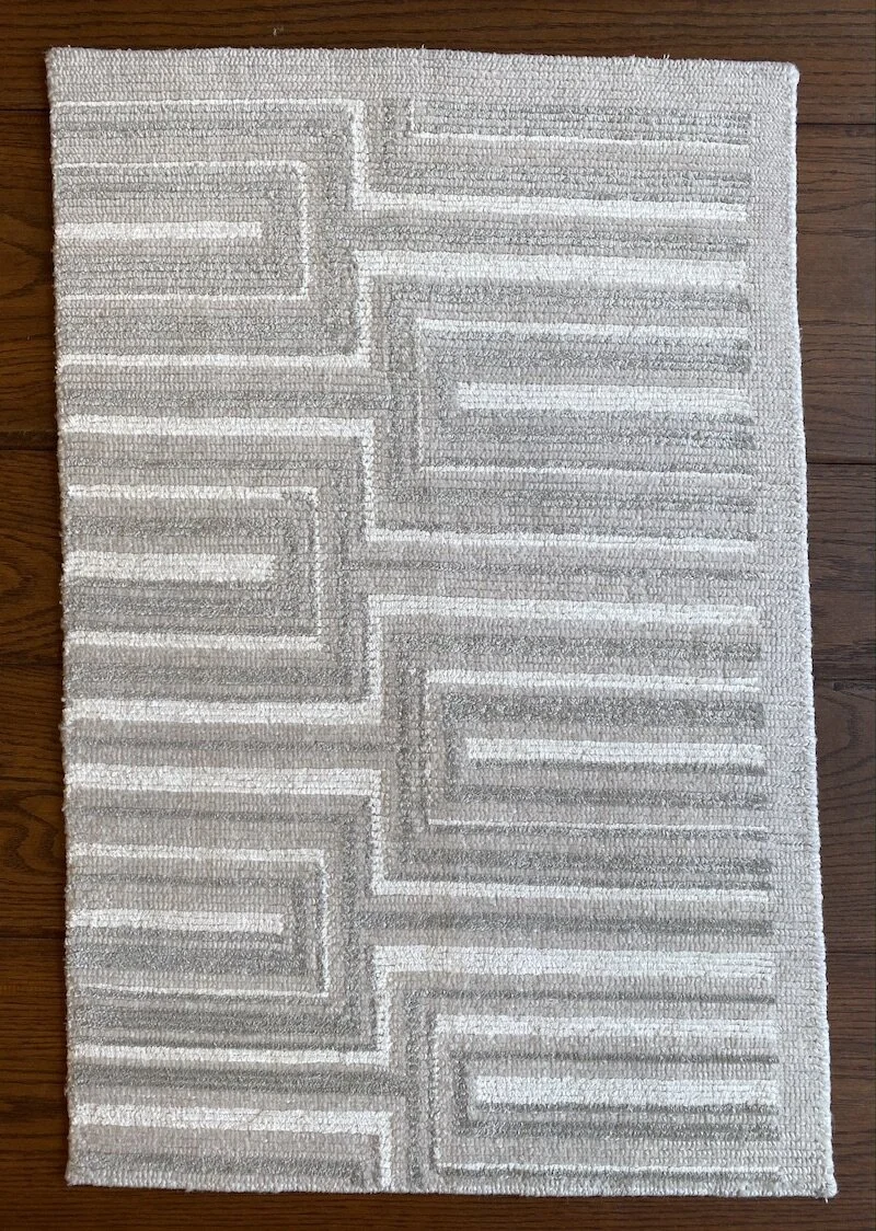 A rectangular area rug with a geometric pattern in shades of white, gray, and beige, placed on a dark wooden floor.