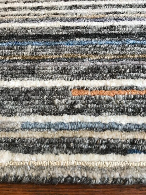 Close-up of a striped carpet with various shades of gray, black, white, tan, and some blue and orange threads.