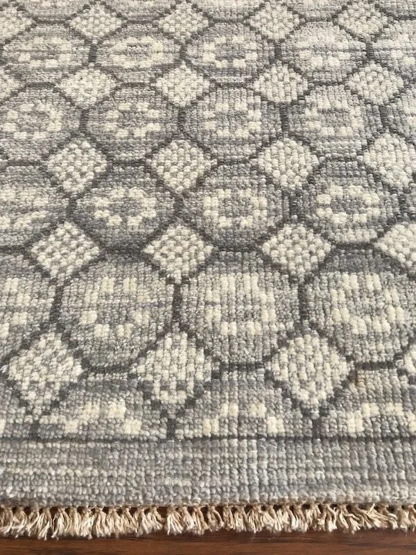 Close-up of a patterned area rug with a geometric design in shades of gray and cream, showing woven texture and fringed edge at the bottom.