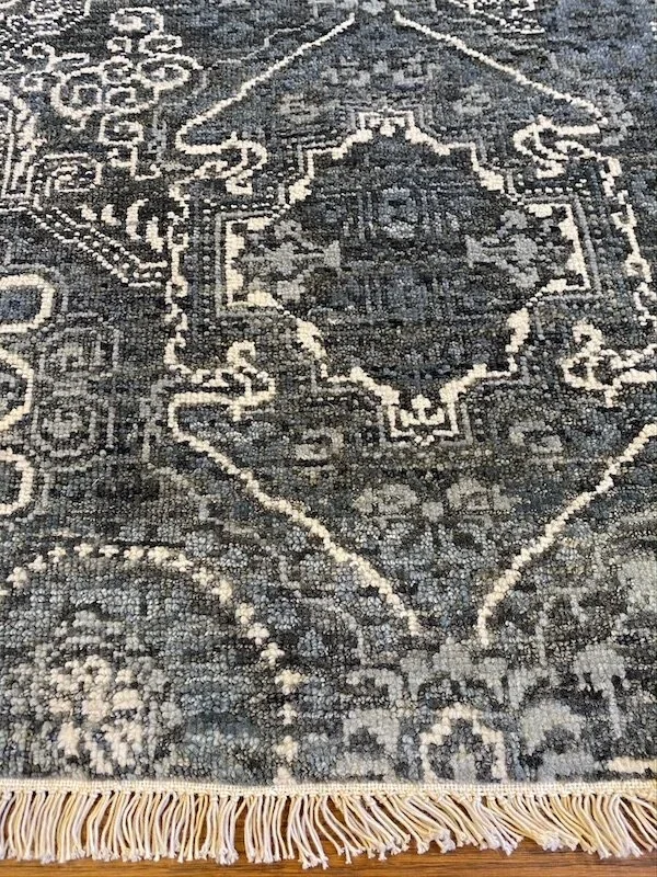 Close-up of a traditional woven area rug with floral and geometric patterns in black, gray, and beige, with fringed edges.