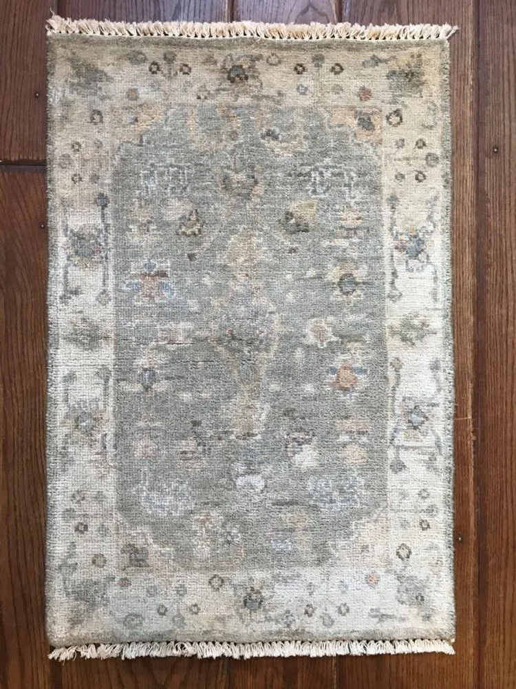 A rectangular area rug with a light, faded design in beige, gray, and muted tones, featuring a central medallion and floral motifs around the edges, placed on a hardwood floor.