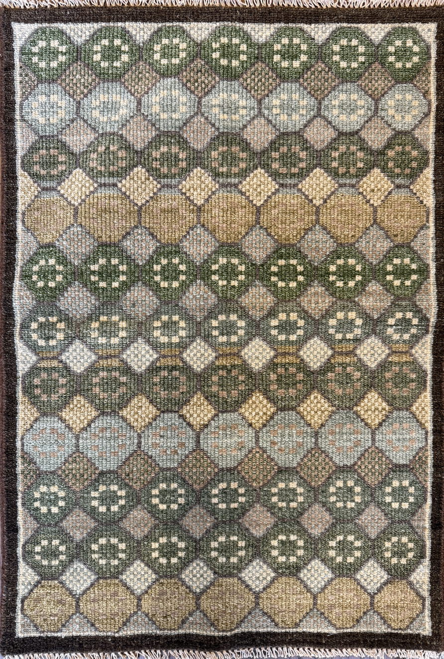 A patterned area rug with geometric designs in green, blue, brown, yellow, and cream colors, bordered by a dark brown frame.
