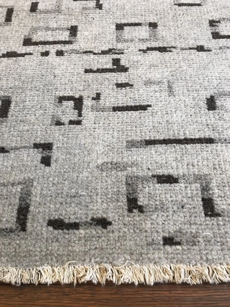 Close-up of a beige and gray patterned area rug on a wooden floor with fringe at one edge.