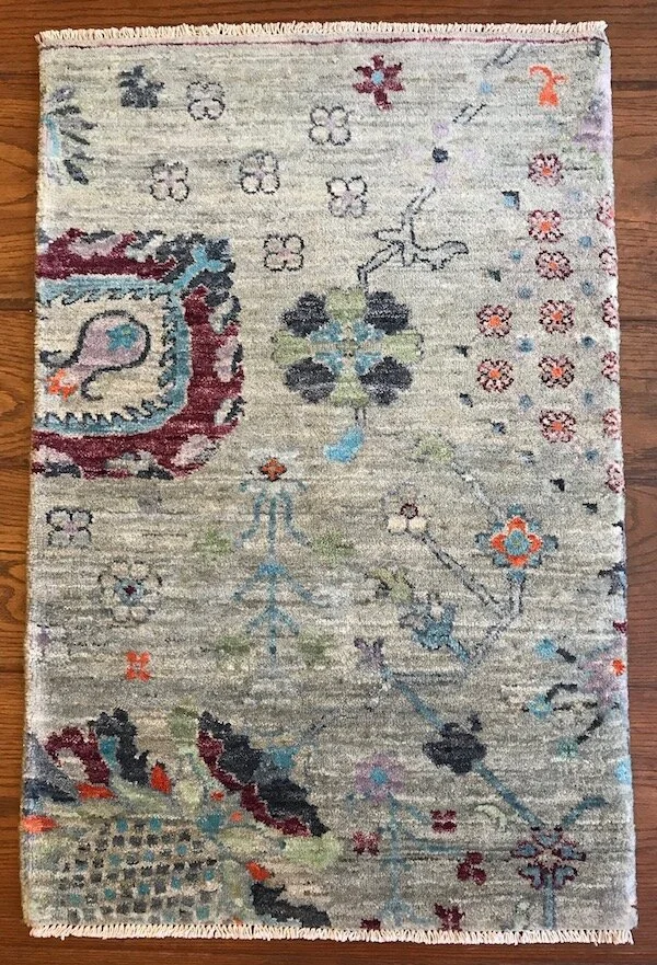 A decorative area rug with floral and abstract patterns in light beige, maroon, black, blue, orange, and green colors, placed on a wooden floor.