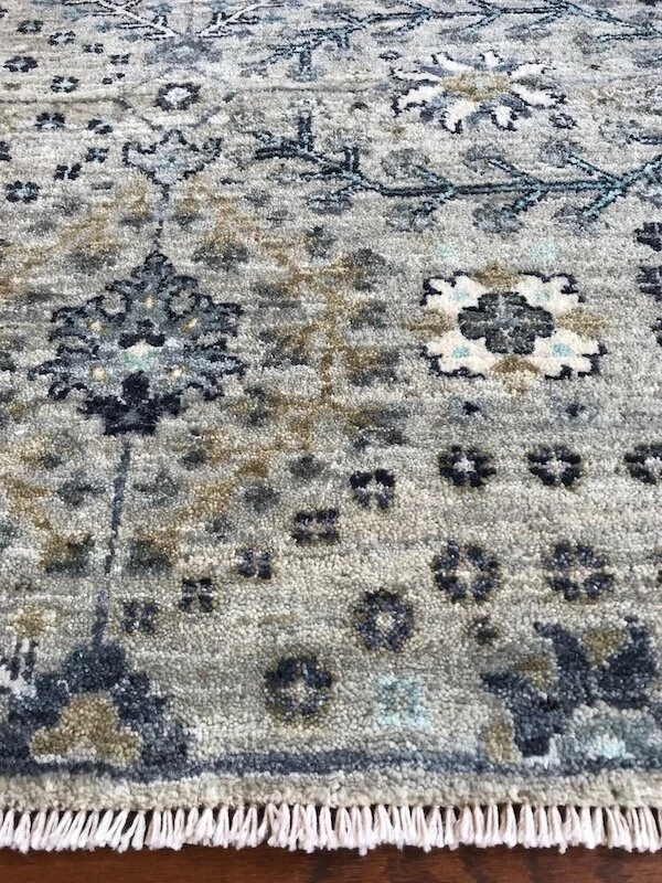 Close-up of a patterned area rug with floral and geometric designs in shades of blue, gray, black, and beige, with fringes at the bottom edge.