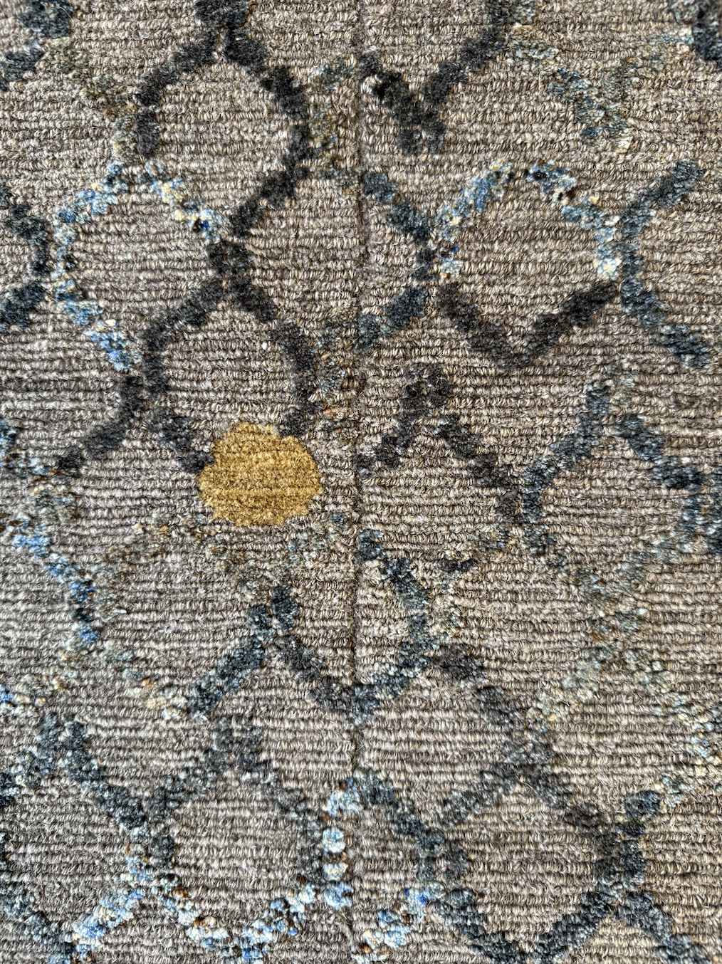 Close-up of a patterned carpet with a floral design in beige, black, and light blue colors, featuring a small yellowish-brown spot.