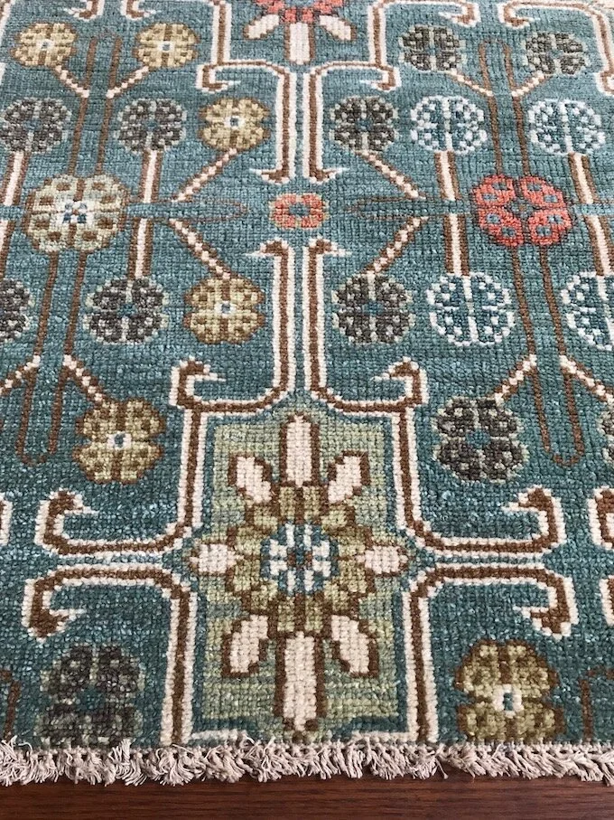 Close-up of a traditional woven area rug with floral and geometric patterns in shades of blue, beige, black, white, and red, with fringed edges.