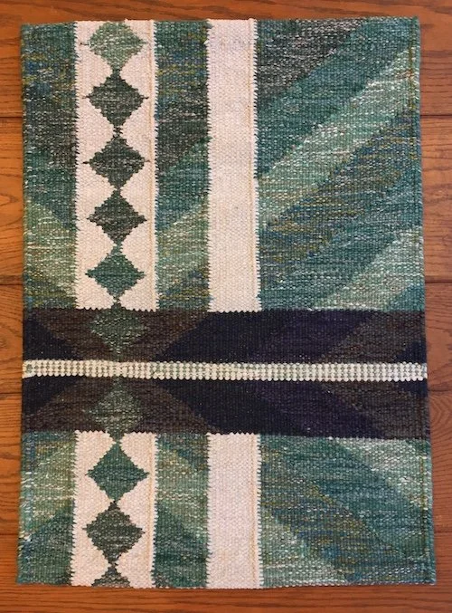 Colorful woven rug with geometric patterns in green, cream, black, and brown tones on a wooden floor.