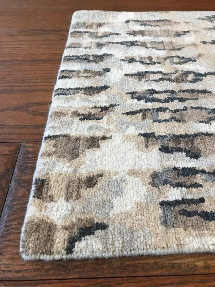 A rectangular area rug with an abstract pattern in shades of beige, brown, gray, and black, placed on a hardwood floor.