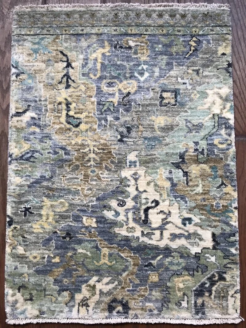 A rectangular area rug with intricate, traditional patterns in shades of blue, beige, and green, placed on a wooden floor.