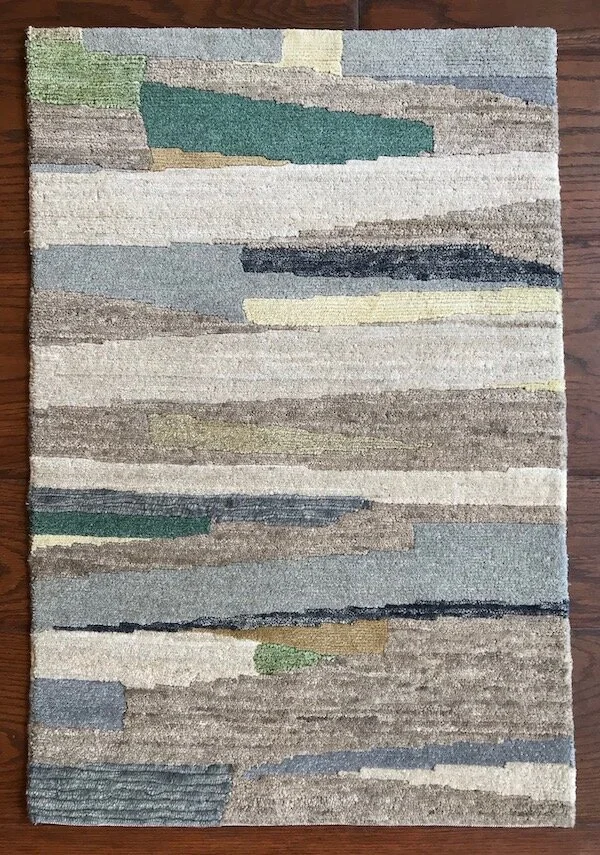 Rectangular area rug with abstract horizontal stripe pattern in shades of green, blue, gray, black, beige, and off-white on a wooden floor.
