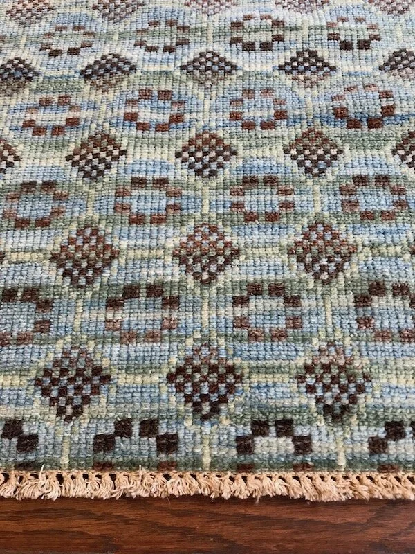 Close-up of a patterned woven area rug with geometric shapes in shades of brown, beige, blue, and green on a hardwood floor.