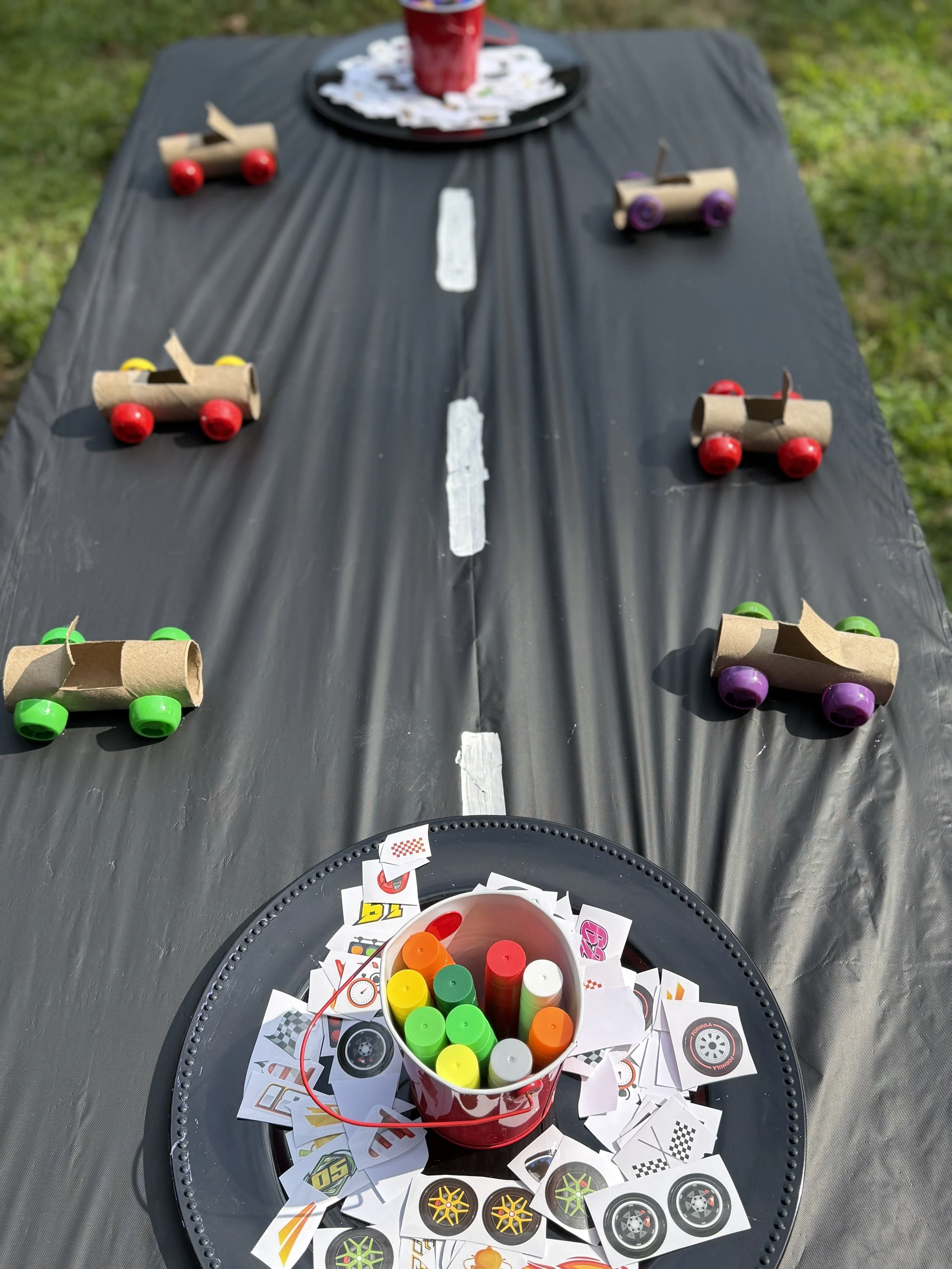 Table decorated for a birthday party with a racing car theme. There are small cardboard cars with colorful wheels, a black tablecloth, a red bucket of colorful markers, and racing-themed stickers scattered on the table. In the background, there is a 