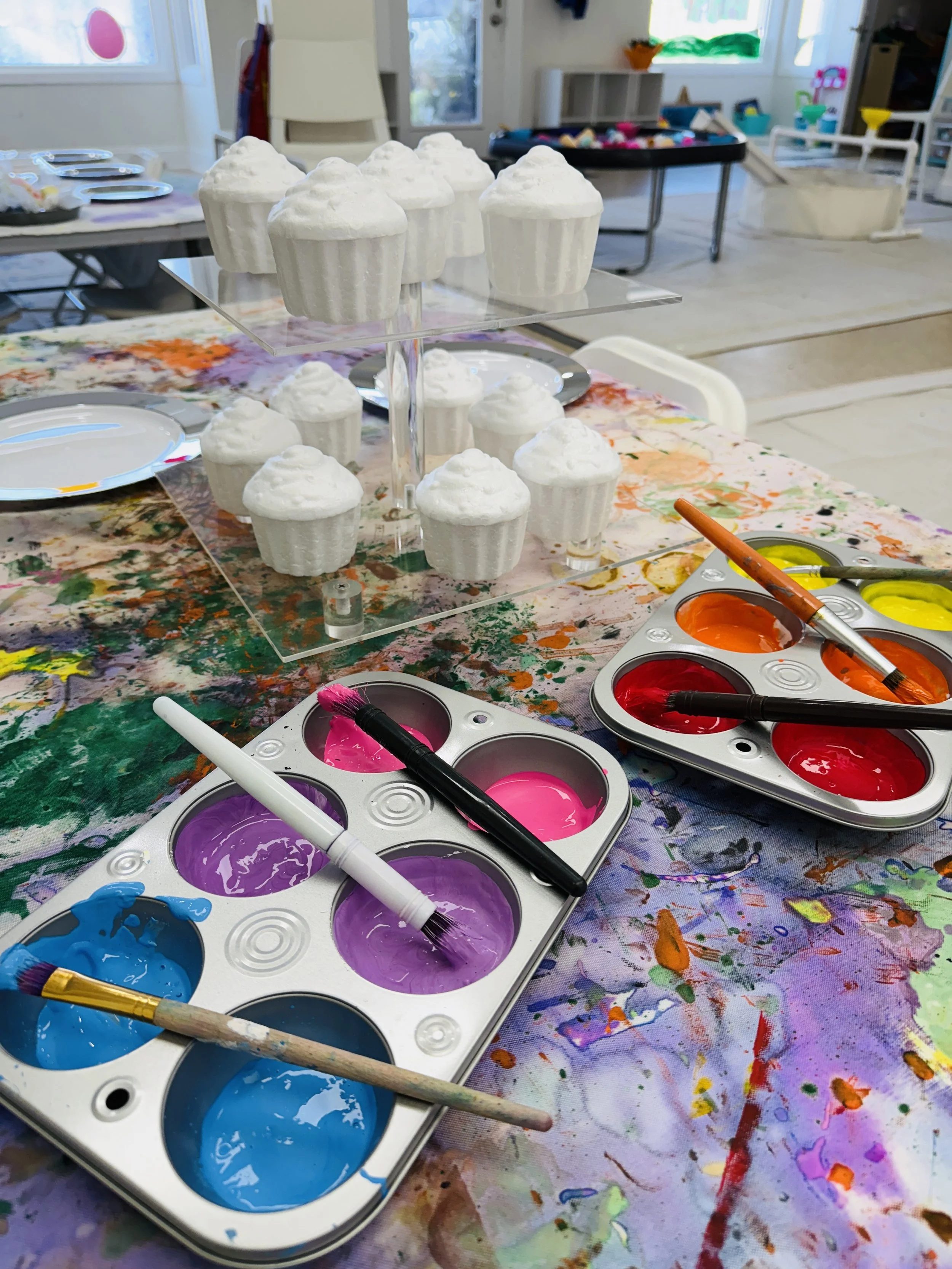 Paint palette with pink, purple, blue, orange, red, and yellow paints, paintbrushes, and children’s toy cupcakes with white frosting on a colorful, paint-splattered table in a bright, playful craft room.