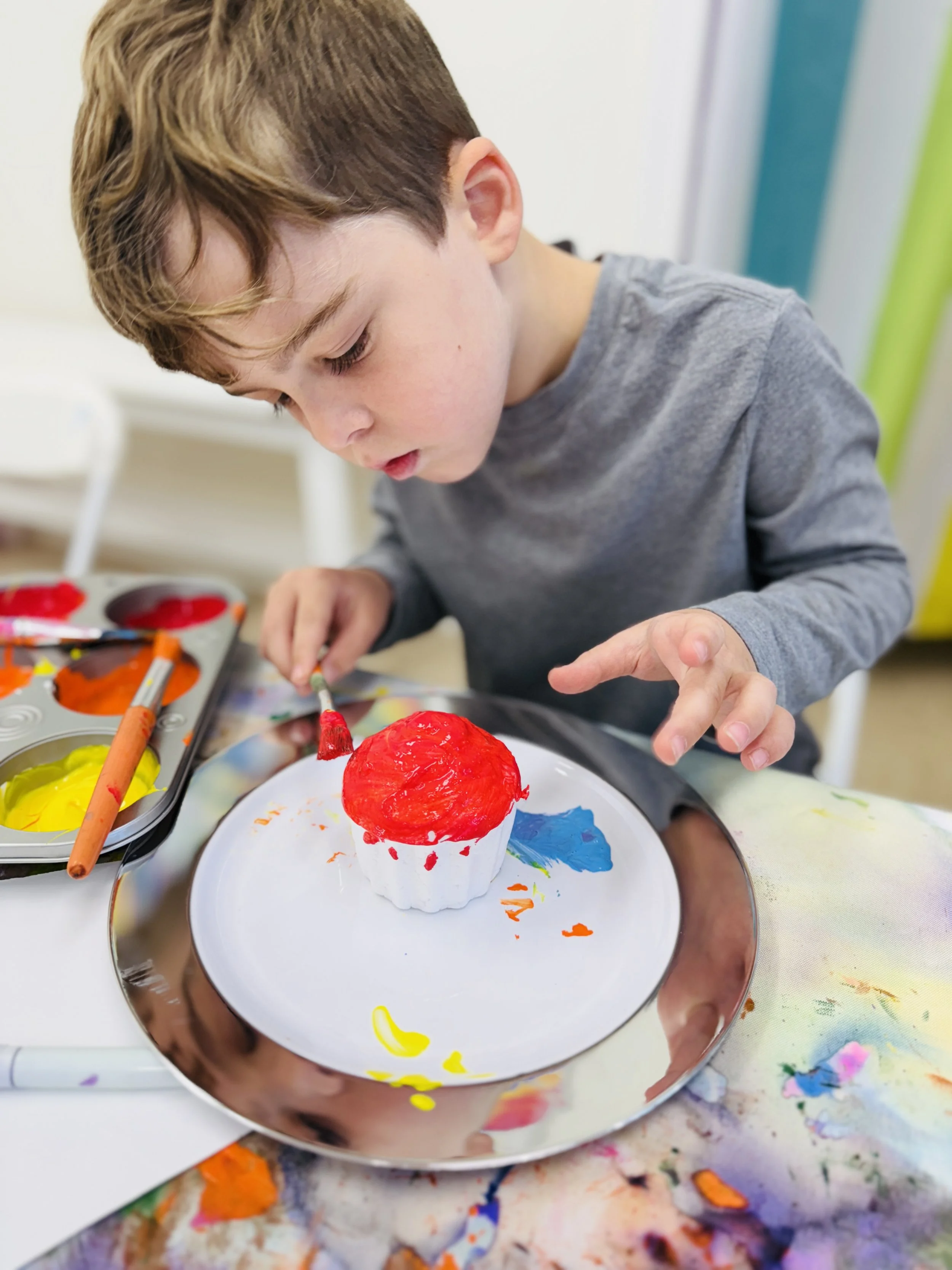 A young boy painting a cupcake with red paint at a table covered with a paint-stained cloth.