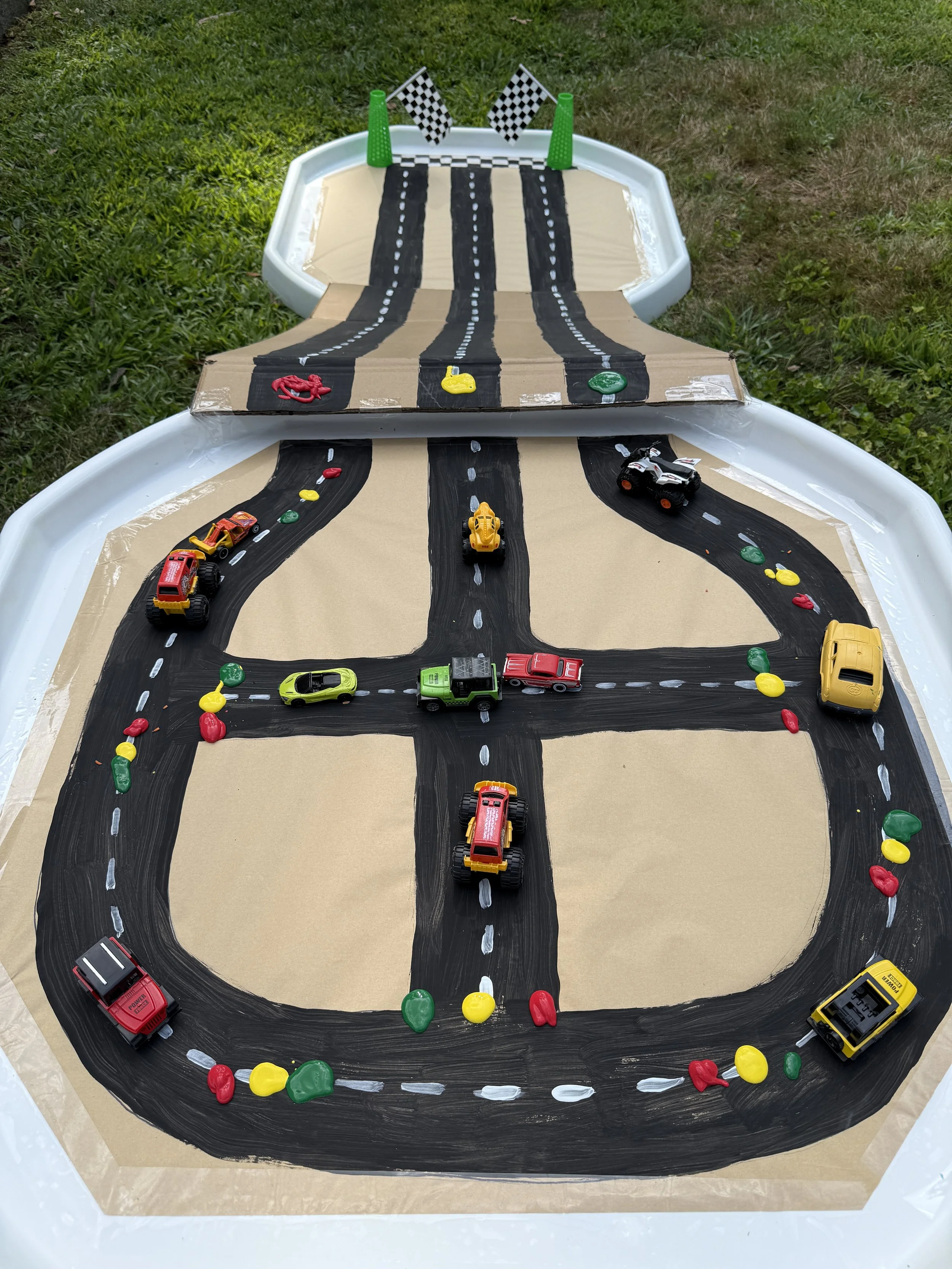 Miniature race track with toy cars, painted roads, and checkered flags on a grassy outdoor surface.