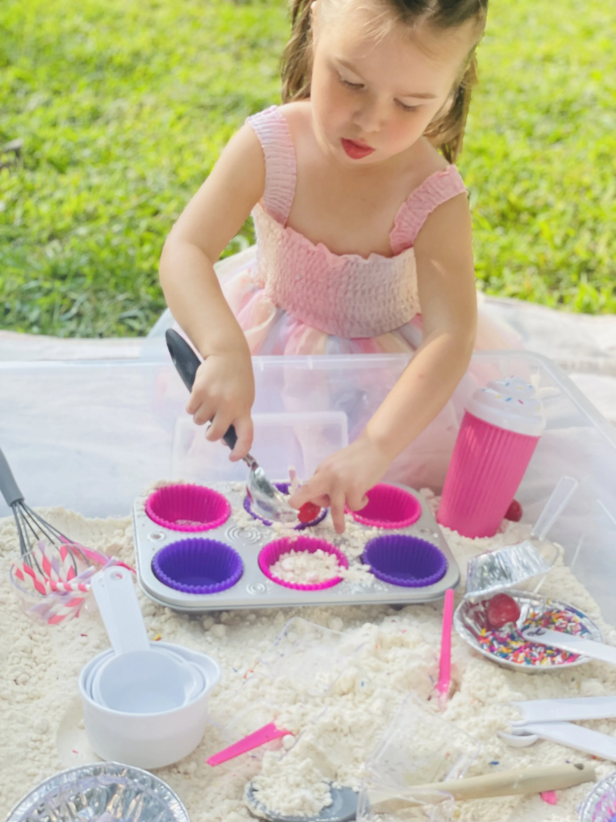 A young girl wearing a pink dress playing with colorful cupcake liners and sprinkles on a table filled with sand, baking utensils, and decorative items outdoors.