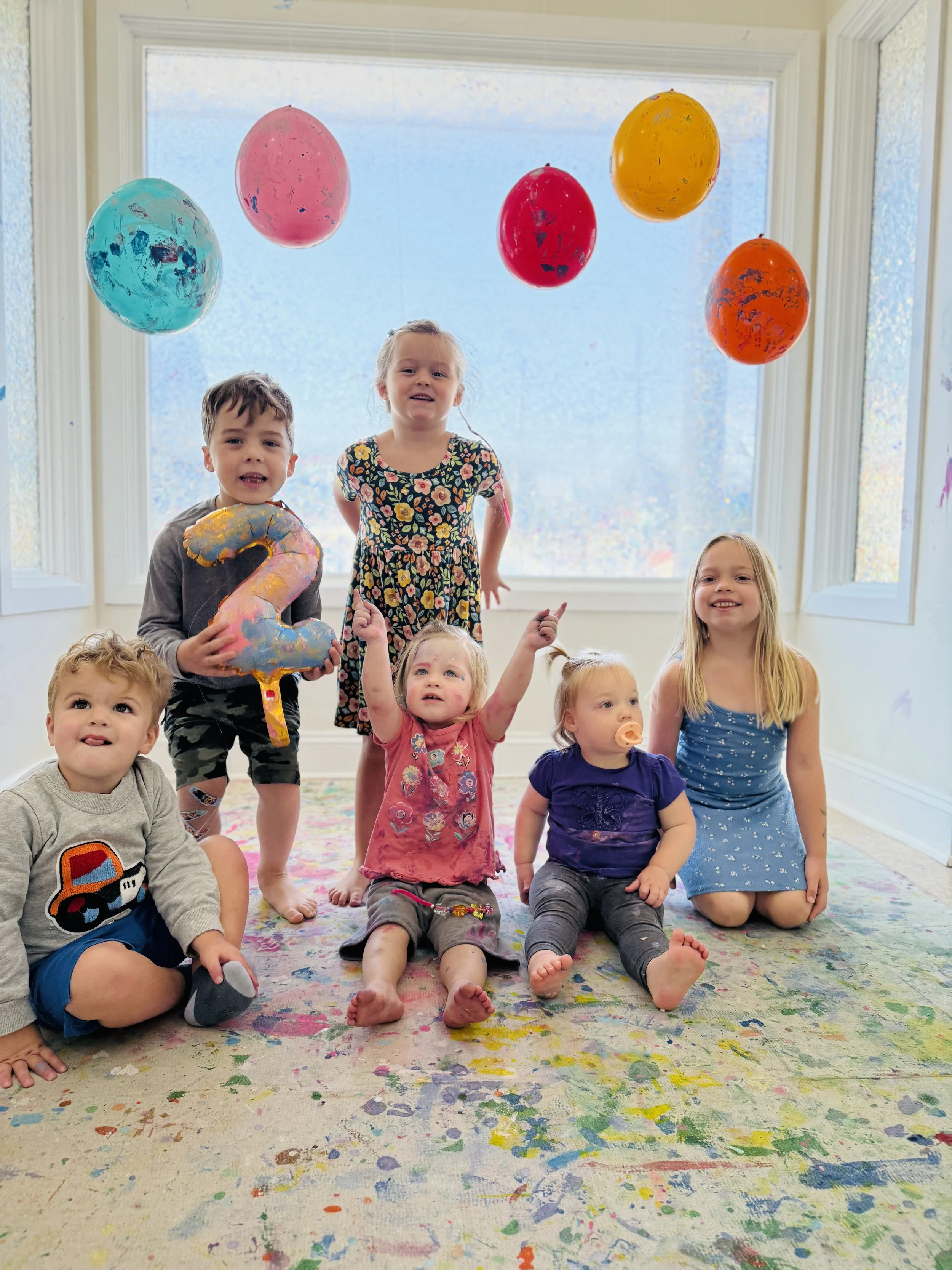 Group of six young children celebrating with colorful balloons and a large number 2 balloon, on a decorated floor with paint splatters, in front of a large window.