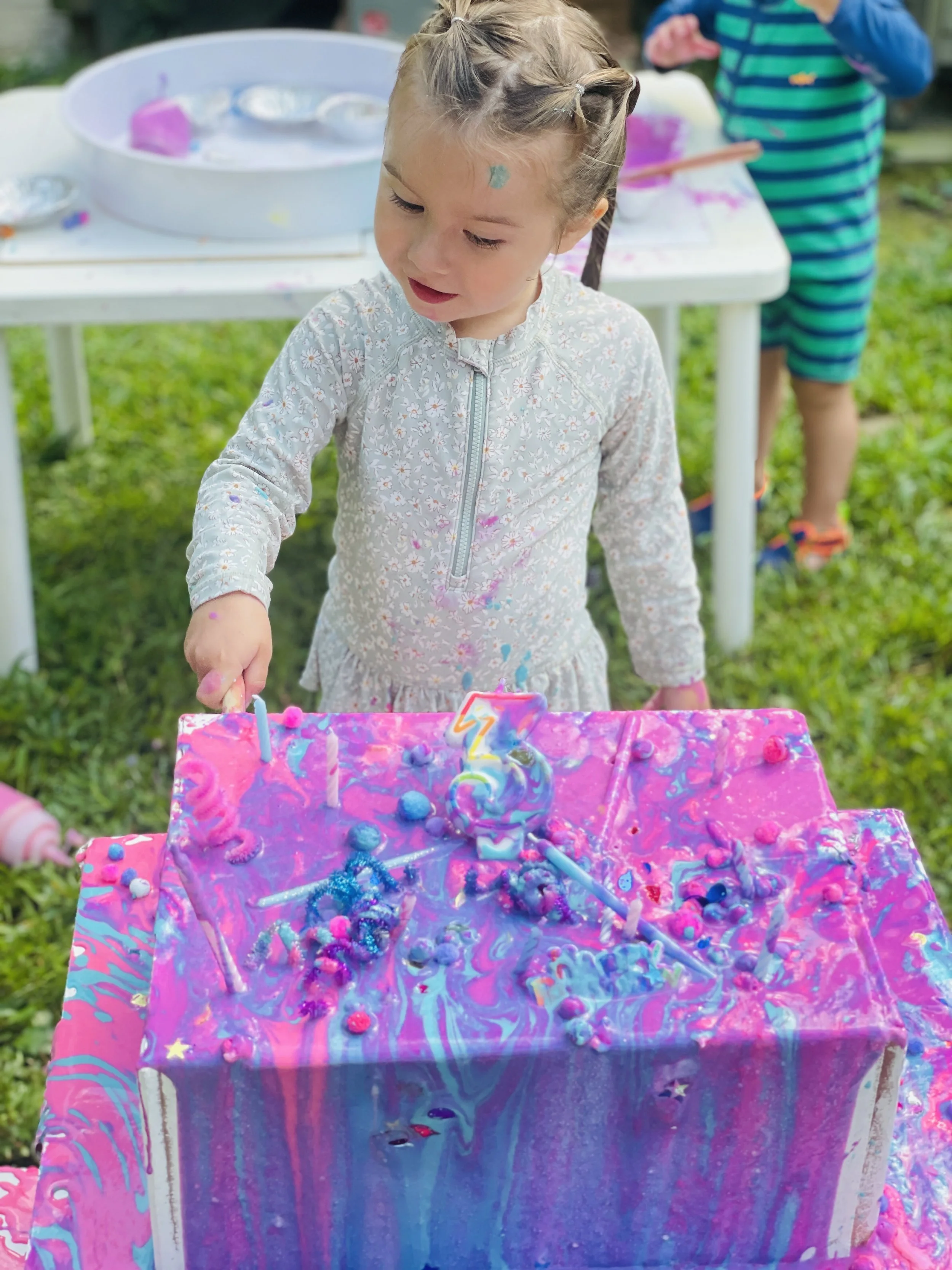 Young girl at a birthday party with a pink and purple marbled cake decorated with birthday candles, sprinkles, and edible decorations, celebrating her third birthday outdoors.