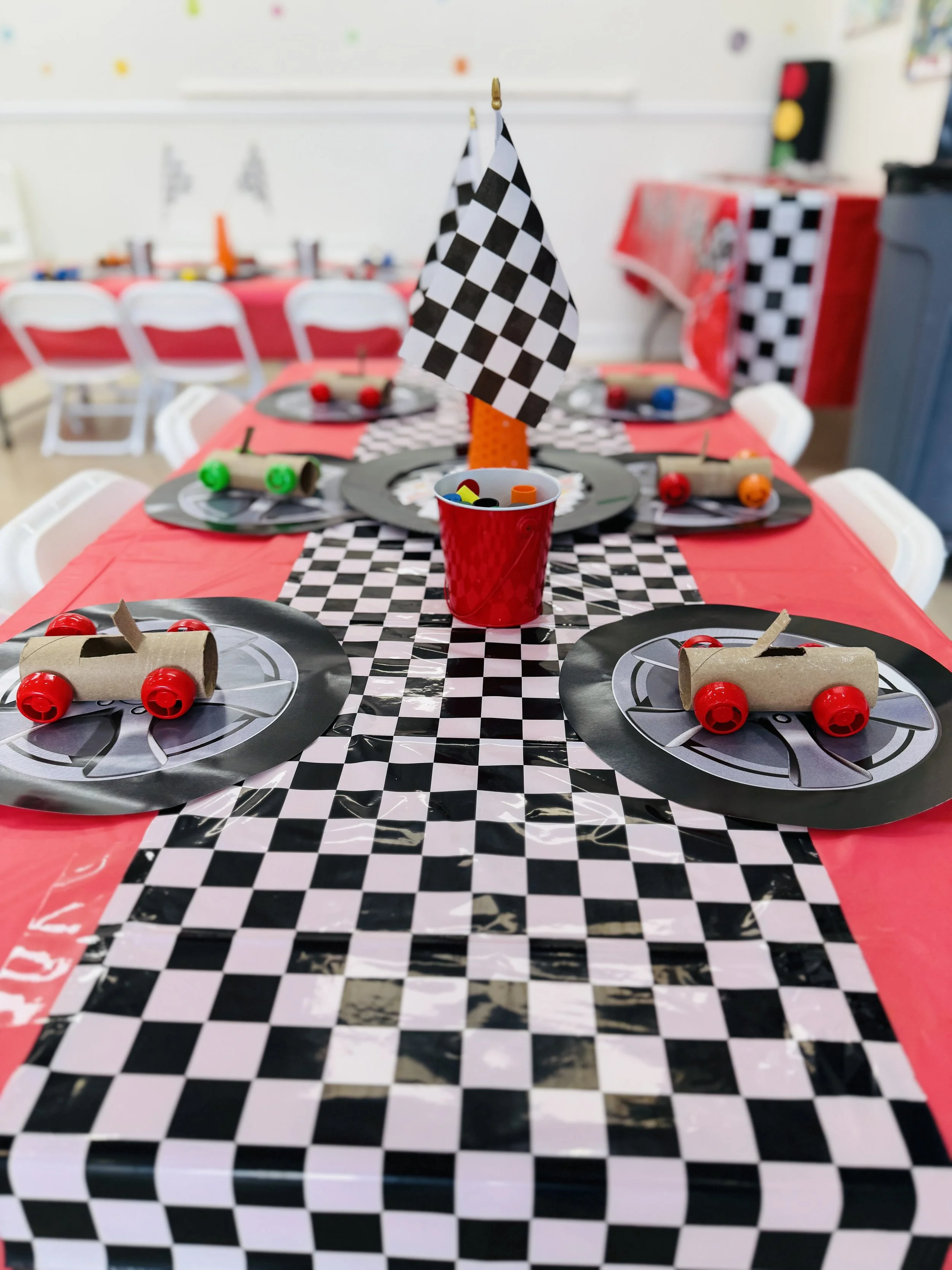 A racetrack-themed party table decorated with a checkered table runner, toy cars, and a checkered flag centerpiece, set in a room with chairs and other decorations.