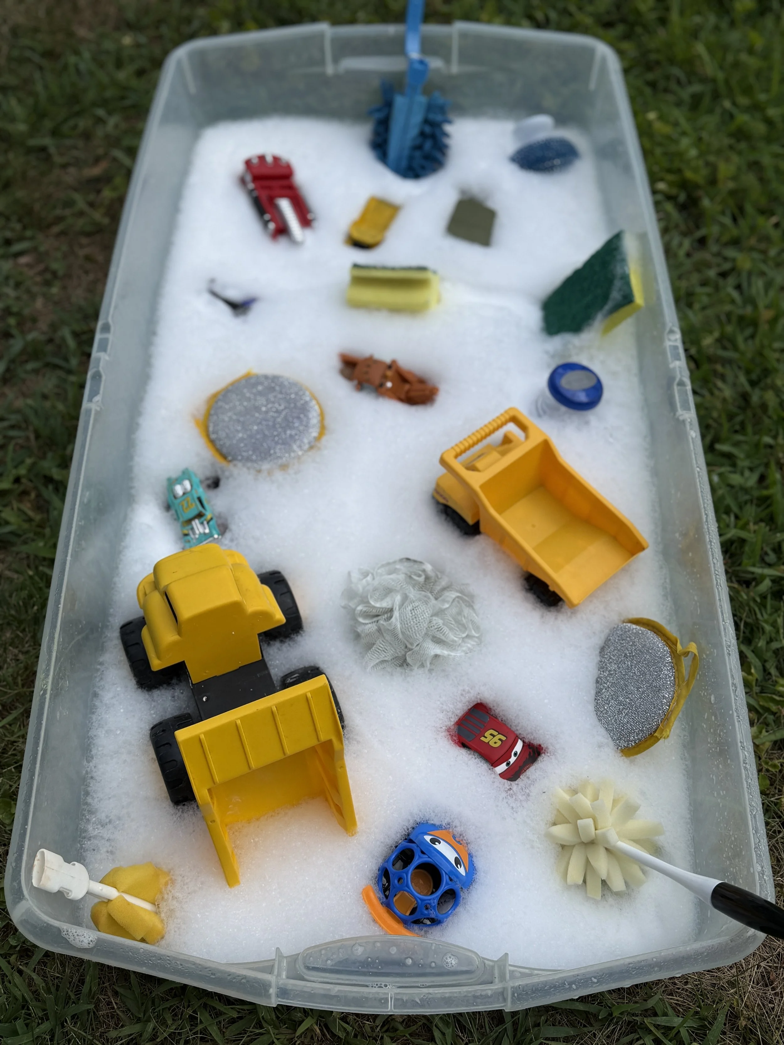 A plastic bin filled with soapy foam containing various toy vehicles and objects, including a red fire truck, yellow dump trucks, blue ball, and sponge brushes, placed on grass.