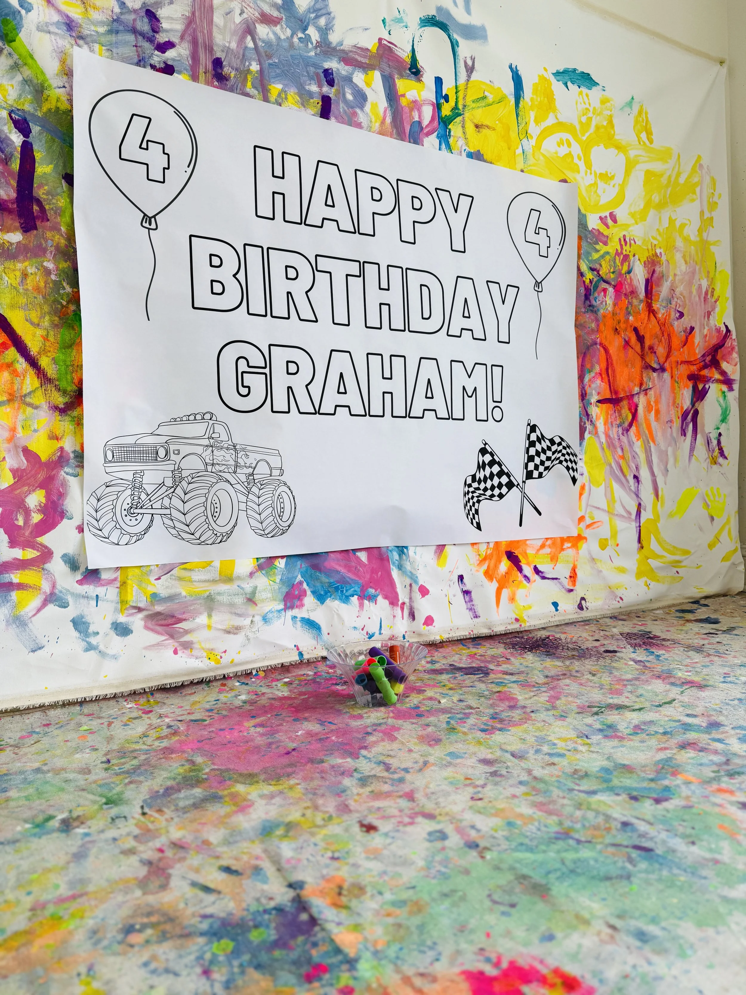 A birthday sign that says "Happy Birthday Graham!" with images of balloons, racing flags, and a monster truck sketch, set against a colorful, abstract painted background and table surface.