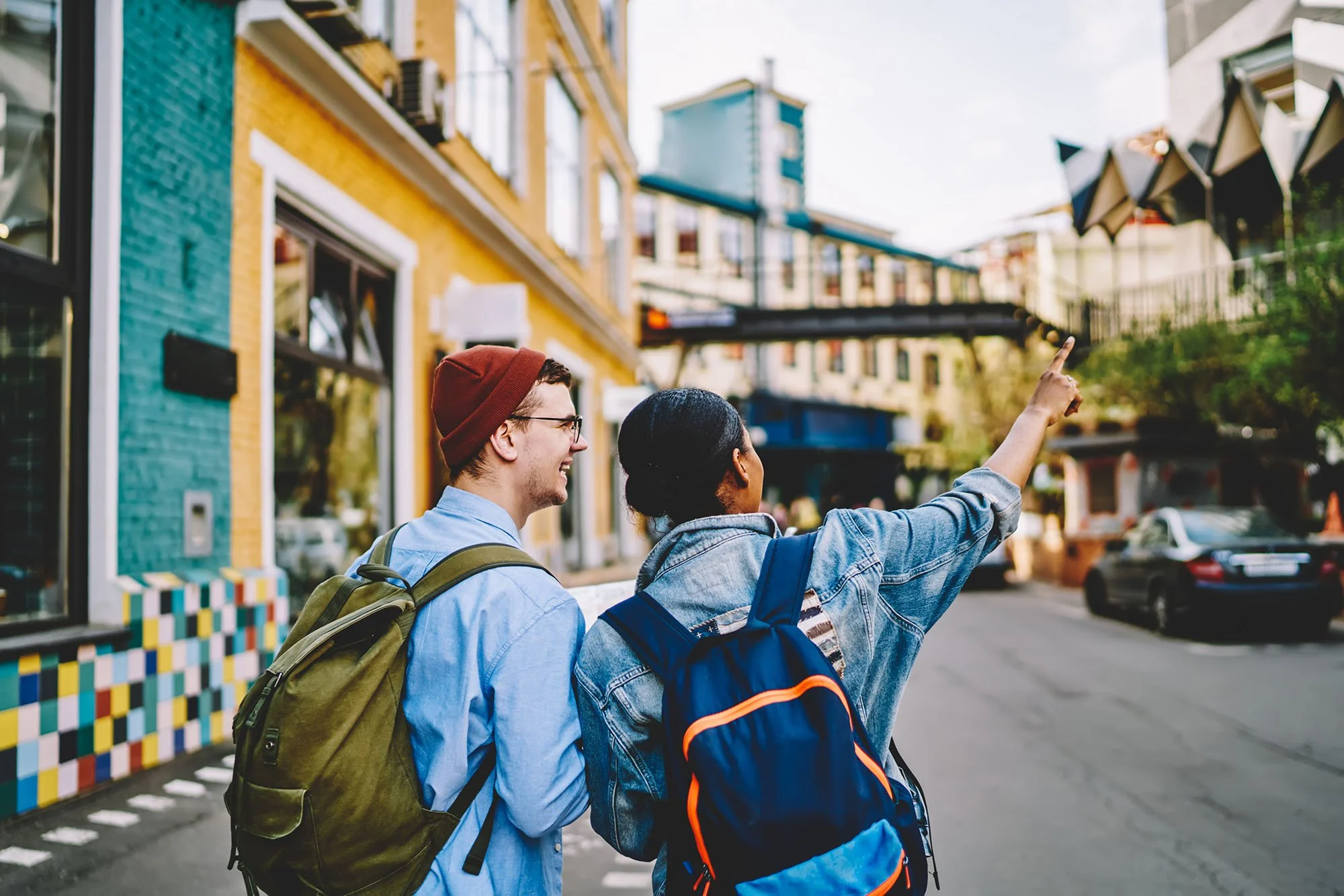 Two young friends with backpacks walking on a city street, one pointing towards a building. Bright colorful buildings and cars in the background.